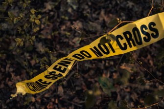 A section of yellow crime scene tape reading “CRIME SCENE DO NOT CROSS” stretches across a wooded area with leaves and branches in the background.