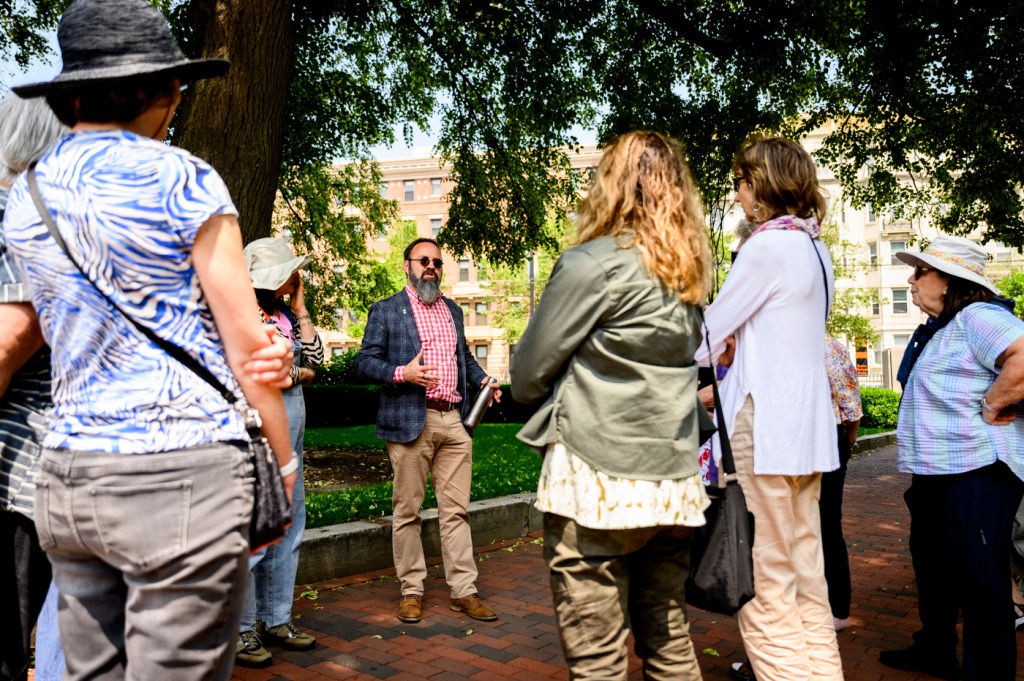 A group of people stands outdoors listening to a speaker during a daytime campus event.
